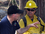 Photograph of two men, one wearing black shirt and cap; another wearing yellow shirt and hard hat.