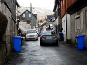 Zwei Pkw fahren auf einer engen Straße zwischen Häusern aneinander vorbei. Zwischen den Autos ist nur wenig Platz. Rechts und links stehen vor mehreren Häusern blaue Mülltonnen.
